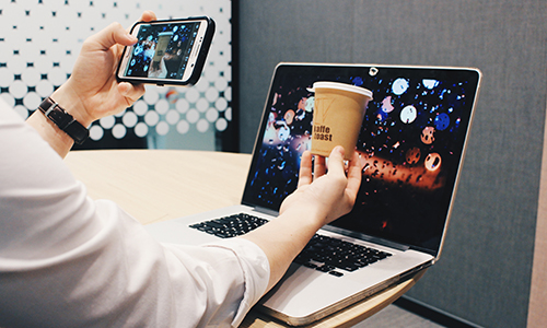 A person taking a picture on a smartphone while holding a cup of coffee in front of a laptop with a bokeh wallpaper as background